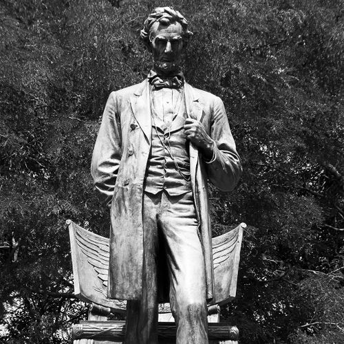 In this celebrated bronze memorial, the figure of President Lincoln stands in a deeply reflective pose before a classically styled chair, from which he appears to have just risen. With his left hand on the lapel of his meticulously rendered period clothing, Lincoln looks slightly downward and steps forward, his left foot just breaking the edge of the plinth on which he stands.