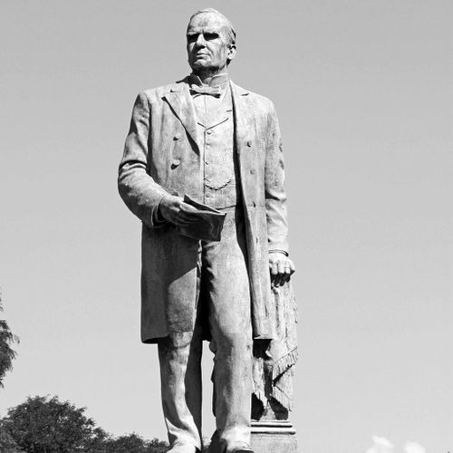 The Mckinley Monument presents an image of the president with his left hand supported on a draped pedestal that stands slightly behind his body, and his right holding a sheaf of papers or folio. Dressed in formal, late 19th-century attire, he looks into the distance in the direction of his right shoulder.