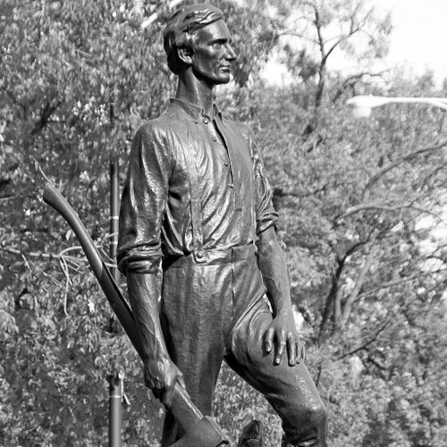 Lincoln, Rail Splitter show the Lincoln as a young man in humble work clothes. Looking resolutely holding an axe at his side in his right hand, his left hand resting on his right leg, which is bent from standing on slightly raised terrain.
