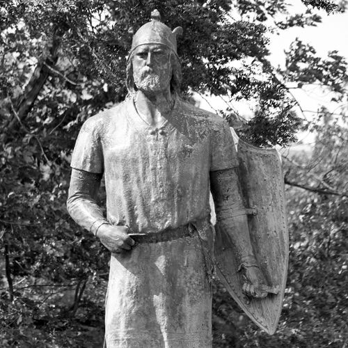 Standing atop a large granite boulder, the bearded figure of Viking Leif Ericson is dressed in period attire, including a helmet and shield that he holds in his left hand. 