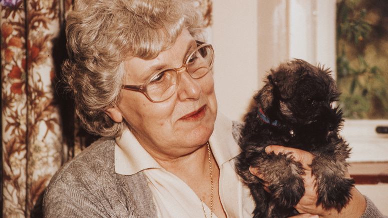 Aged care costs- photo of an older lady with curly grey hair and grey cardigan holding a small brown Maltese type puppy