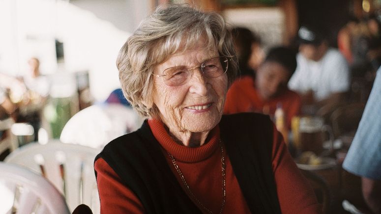 Photo of an older lady, sitting at a cafe while in respite, wearing a rust coloured polo neck with black vest smiling at someone off camera