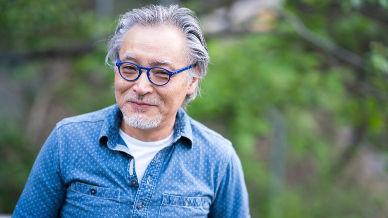 Transitional care- photo of an asian man with grey hair smiling at the camera in a blue shirt and blue round glasses