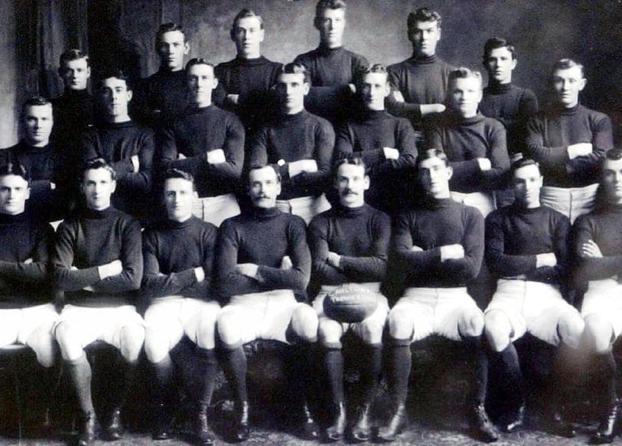Black and white photo of a men's football team wearing black skivvies, white shorts and knee high socks with football boots