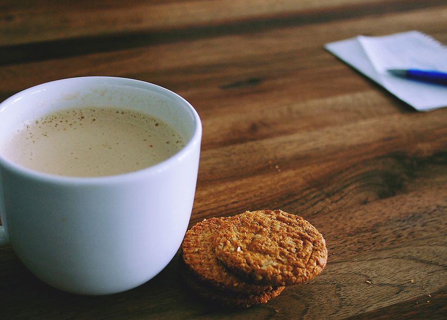 Hacks to get you out of the house- Cropped photo of a cup of tea with two anzac biscuits and a note pad and pen on a wooden table