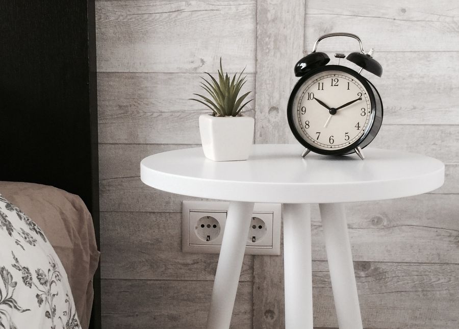 Keep your mind sharp at home- photo of a white round bedside table with alarm clock and plant. 