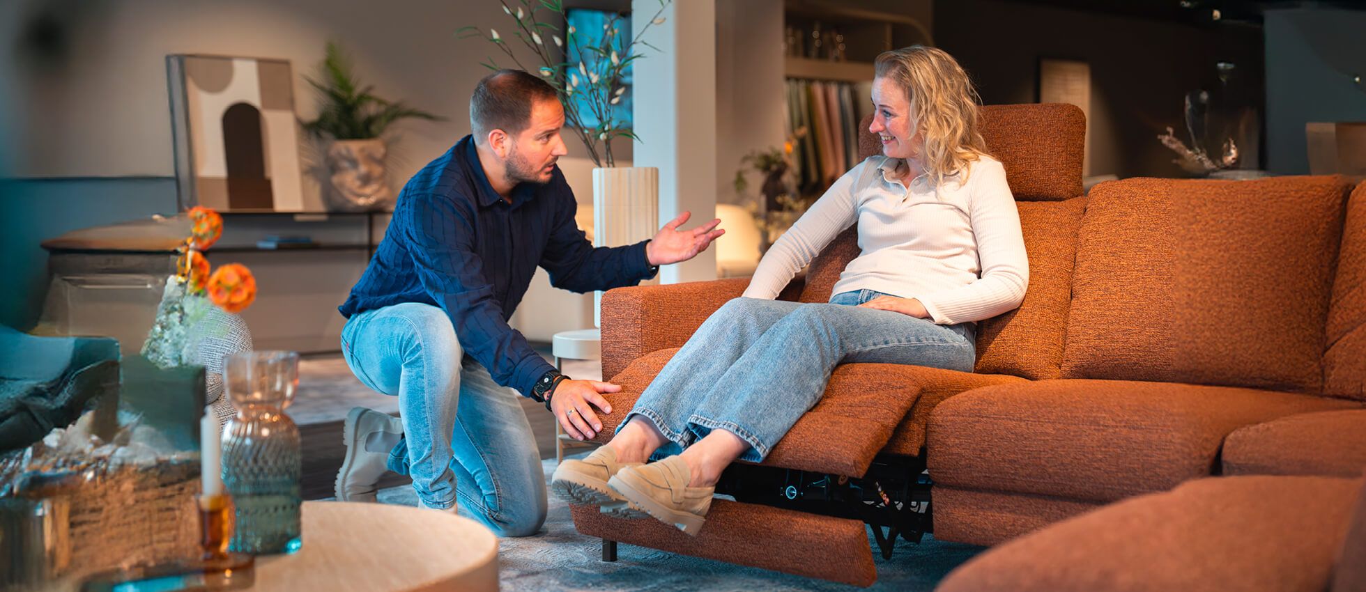 A woman sitting on an orange recliner smiles at a man kneeling beside her, discussing the chair in a modern living room setting.