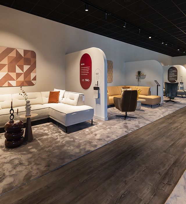 Modern furniture showroom with sectional sofas, armchairs, decorative rugs, and wall art under spotlighting.