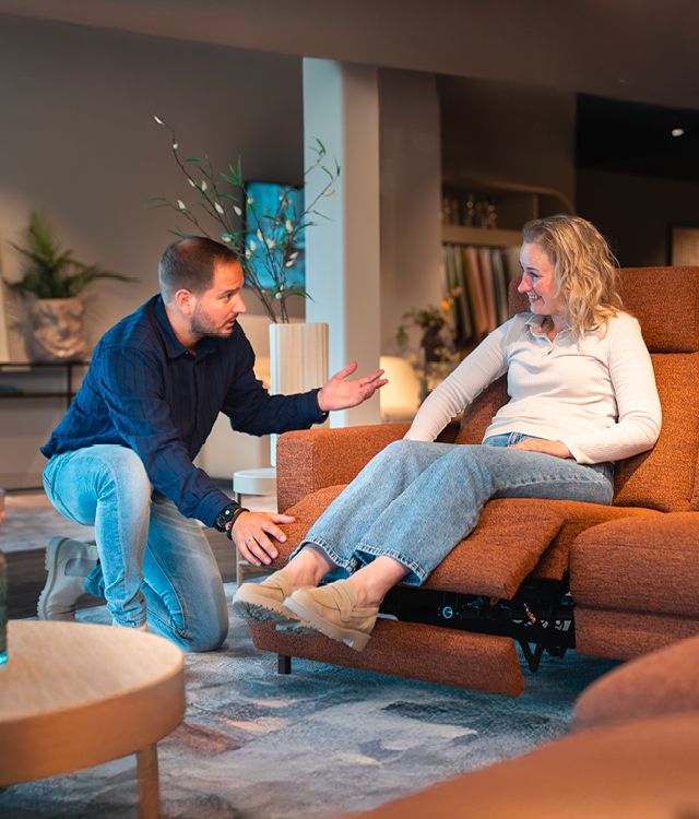 Man kneeling beside a woman sitting on an orange recliner, demonstrating its features in a cozy, modern living room setting.