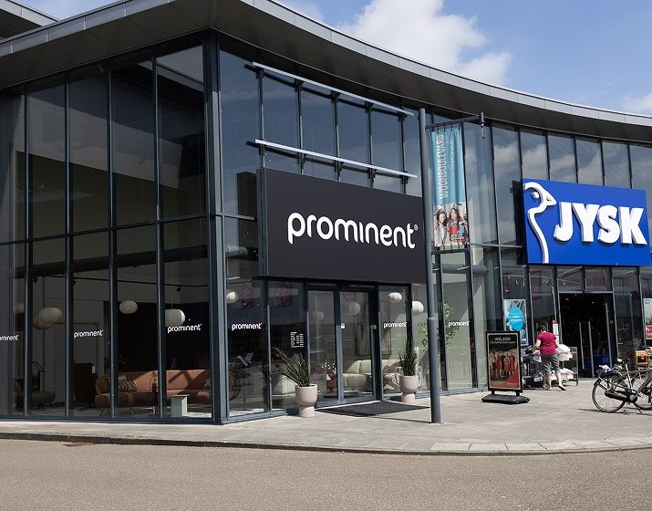 Glass-fronted stores with signs reading "Prominent" and "JYSK" on a curved building, with potted plants and a bicycle rack nearby.