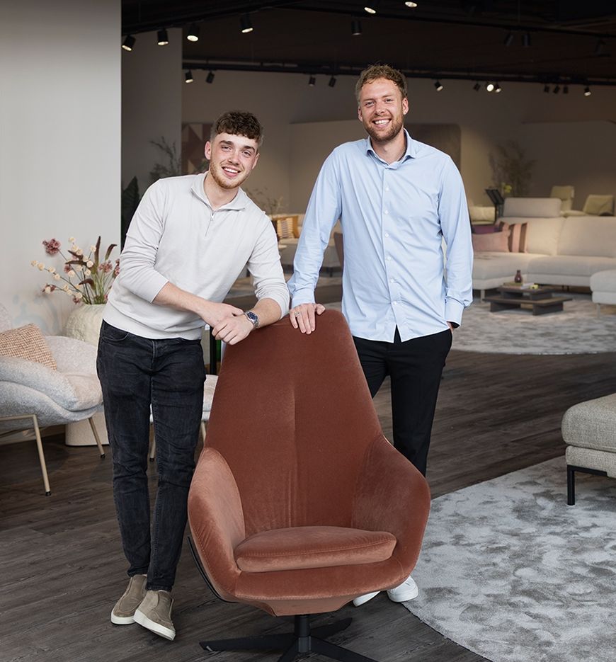 Two men smiling while standing beside a brown velvet swivel chair in a modern living room showroom.