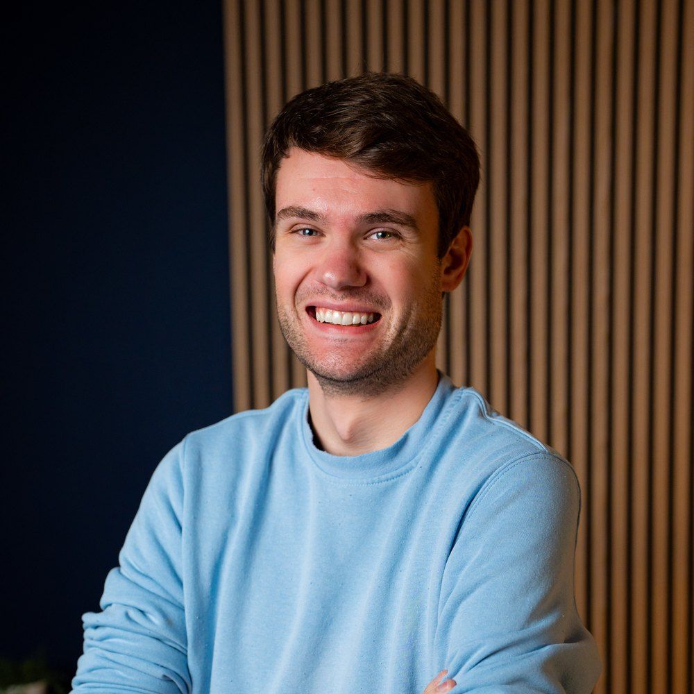 Smiling person with short hair in a brown sweater stands against a wooden wall with small plants on shelves.