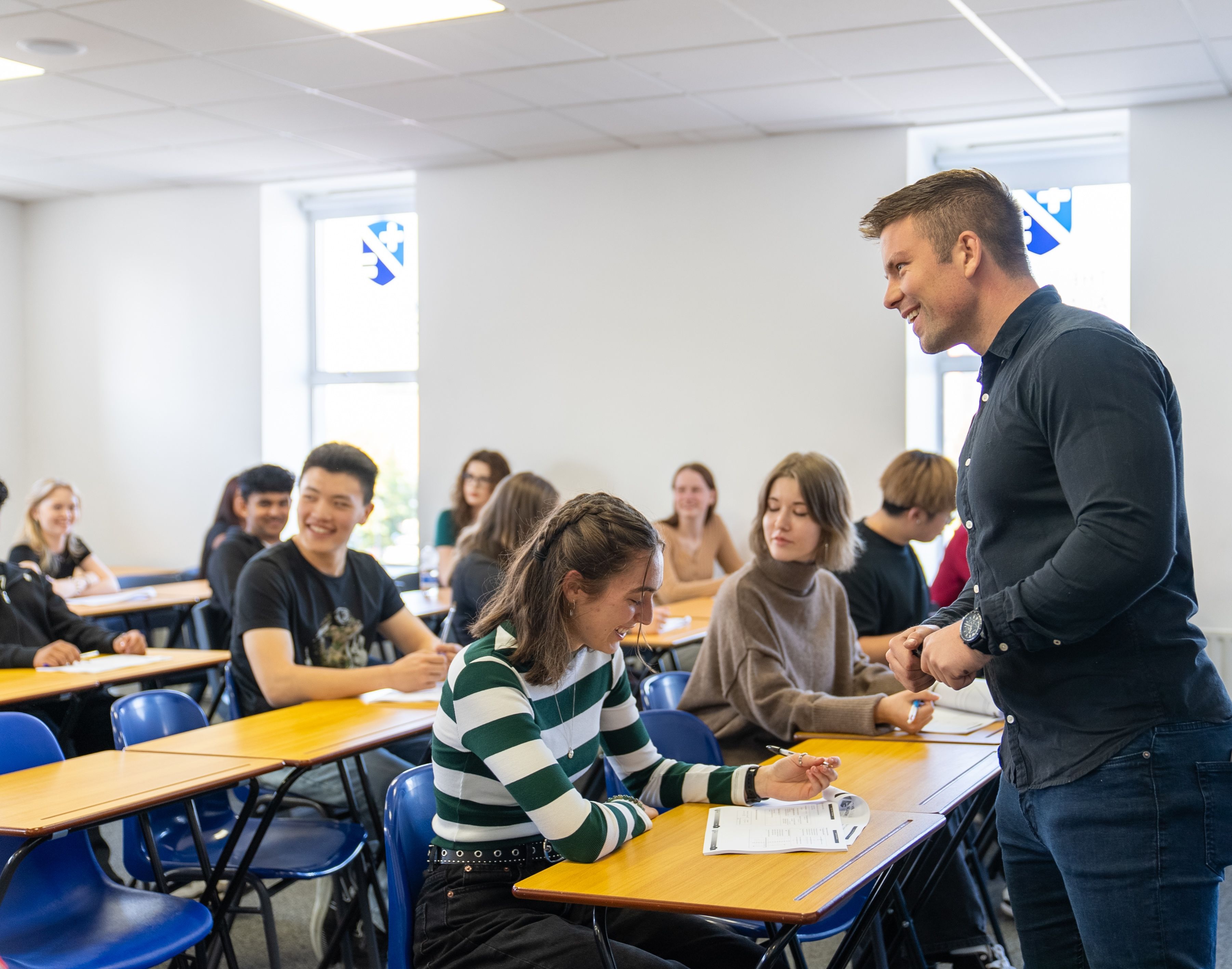 A teacher interacts with students seated at desks in a bright classroom. The students are engaged and taking notes.