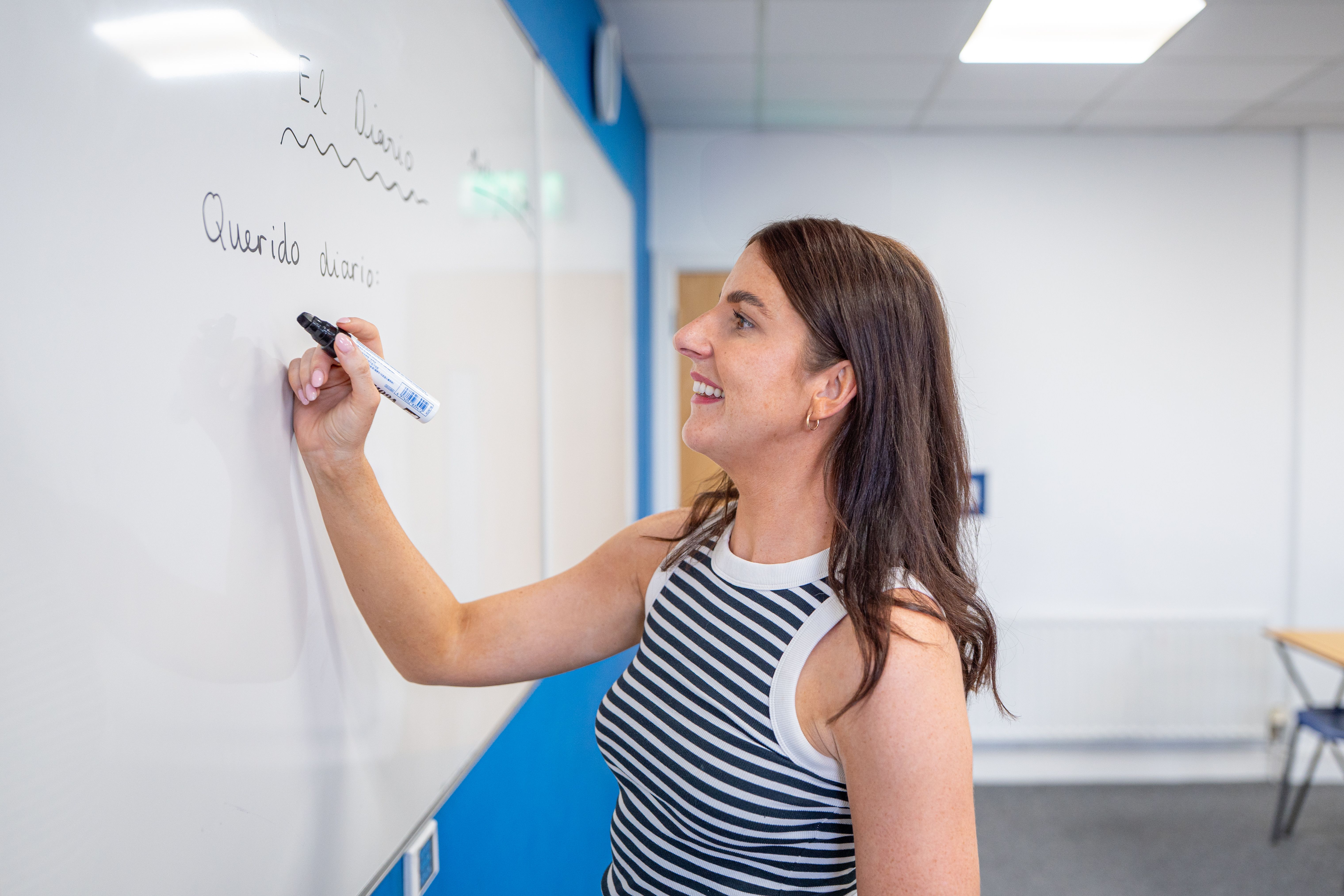 Woman smiling and writing on a whiteboard in a classroom setting, wearing a striped sleeveless top.
