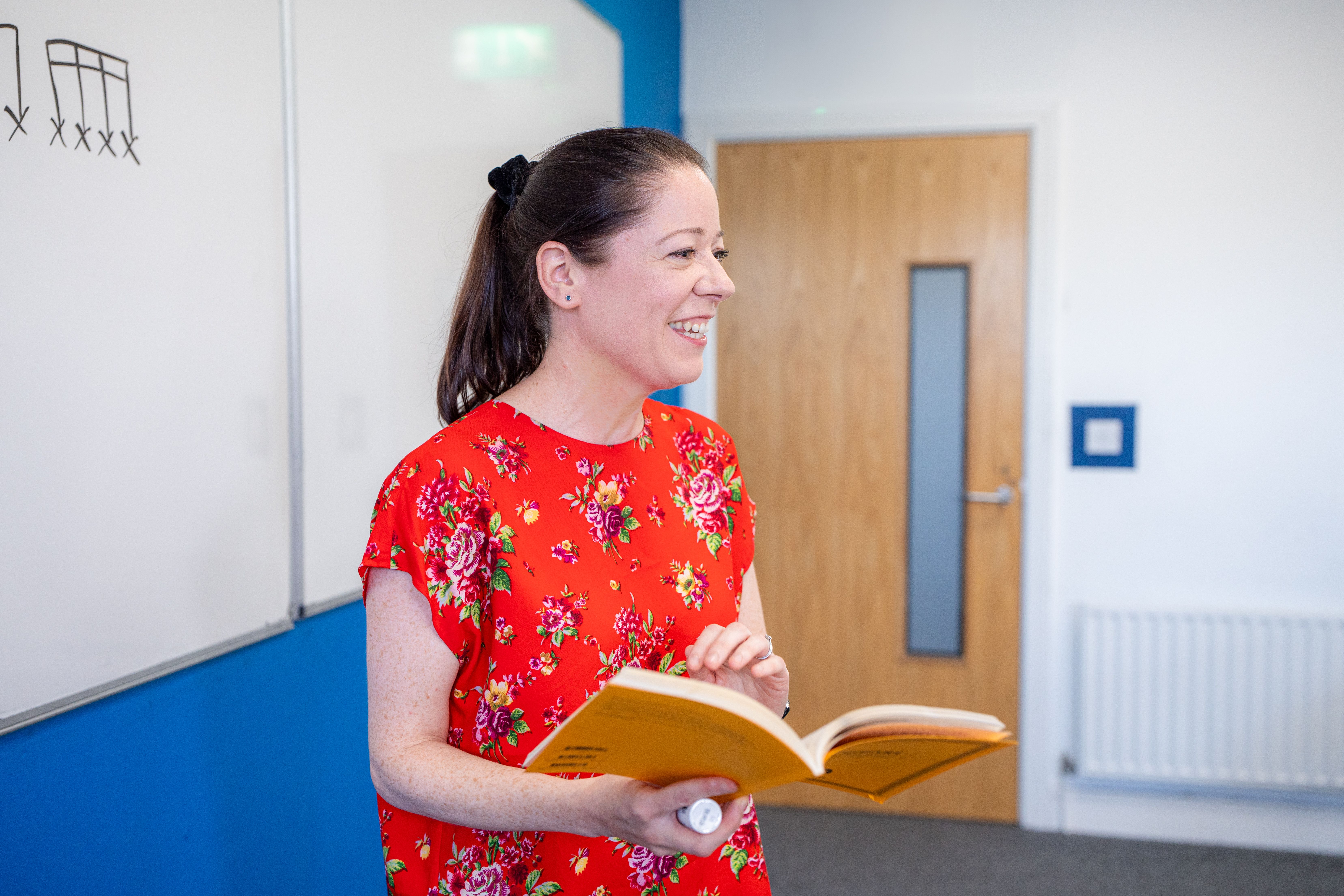 A woman in a red floral dress holds an open book, smiling in a classroom with a whiteboard and diagrams behind her.