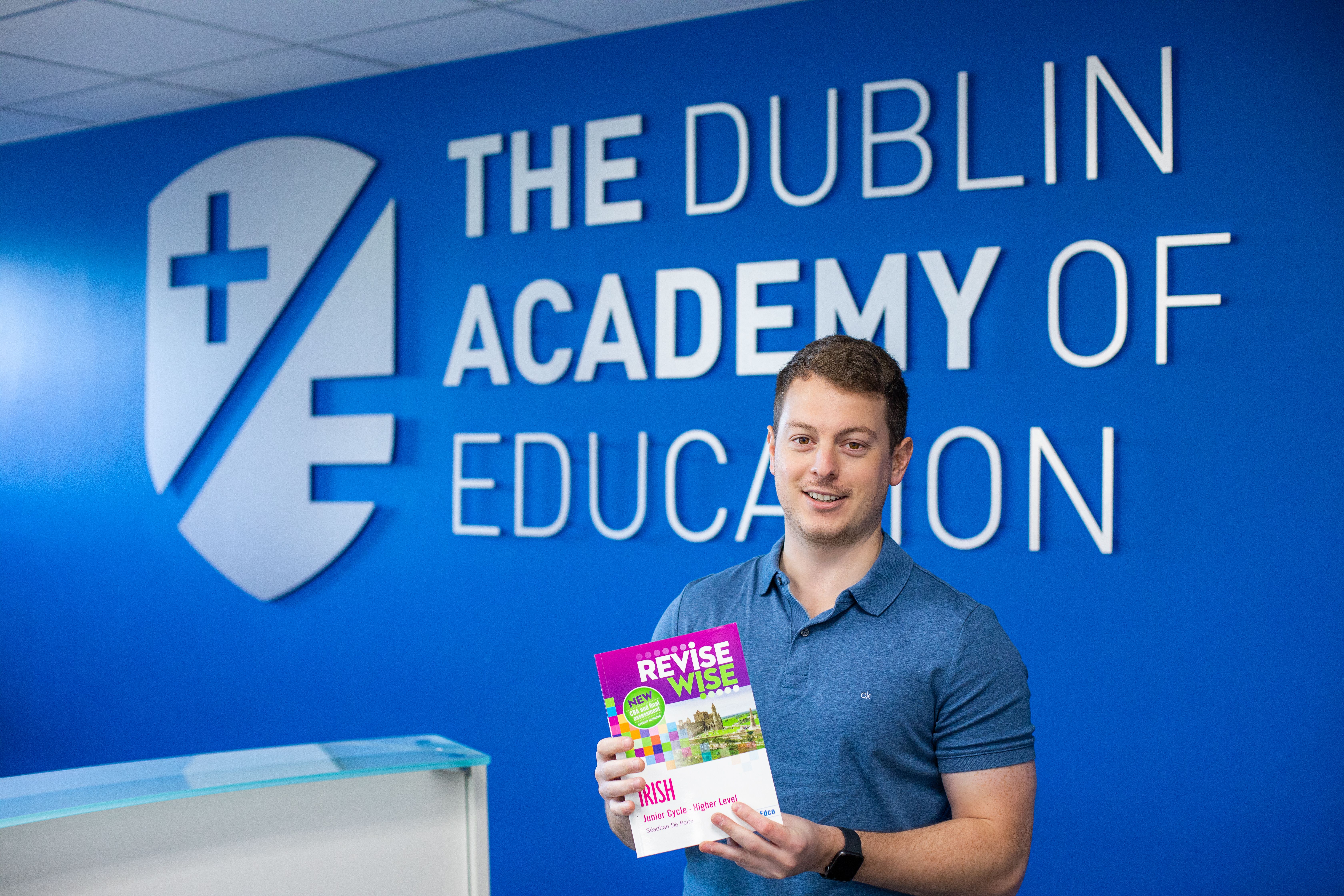 A person in a blue shirt holds a book titled "Revise Wise" in front of a blue wall with "The Dublin Academy of Education" logo and text.