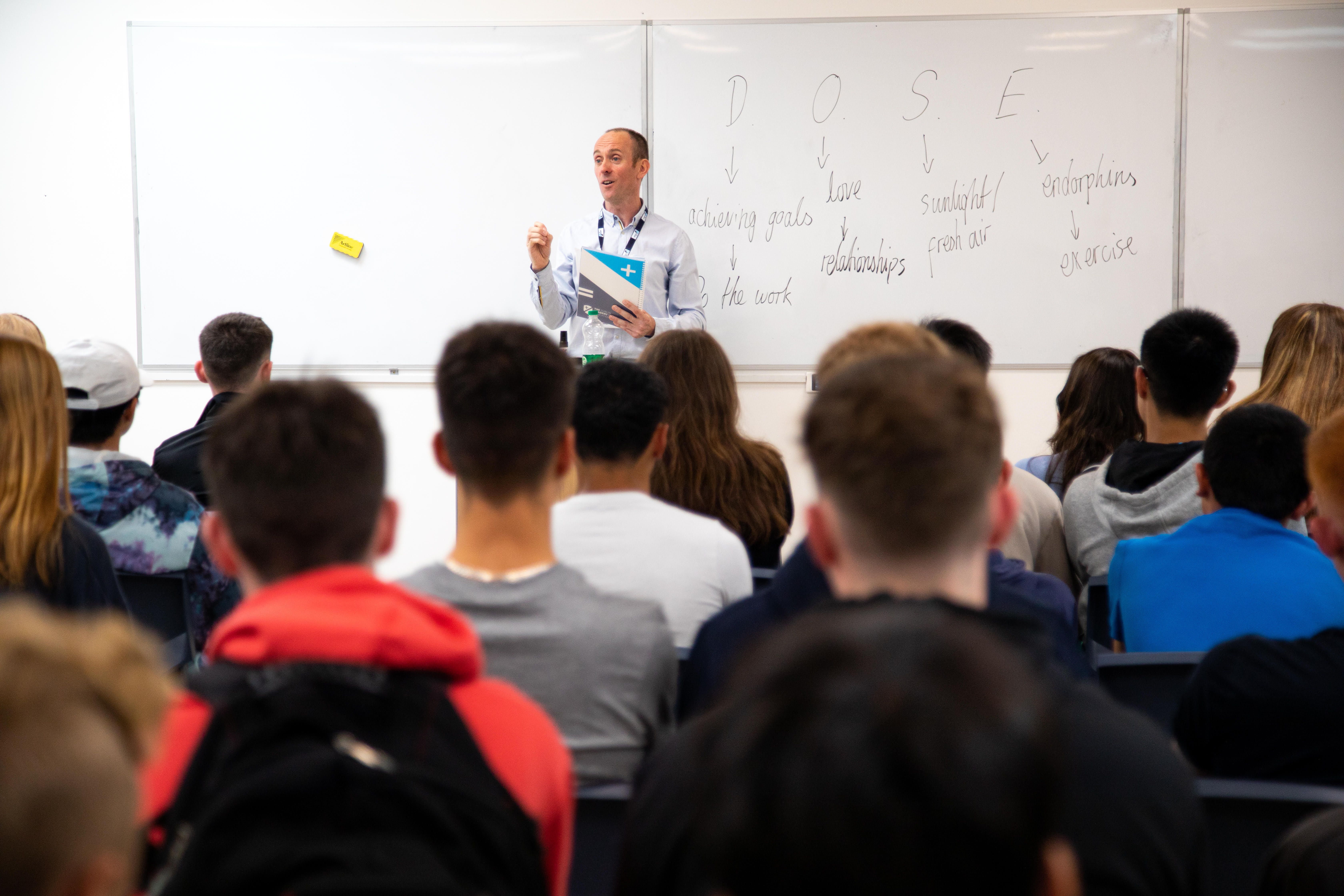 A teacher lectures to a classroom of students, with notes on a whiteboard labeled "DOSE" and various words.