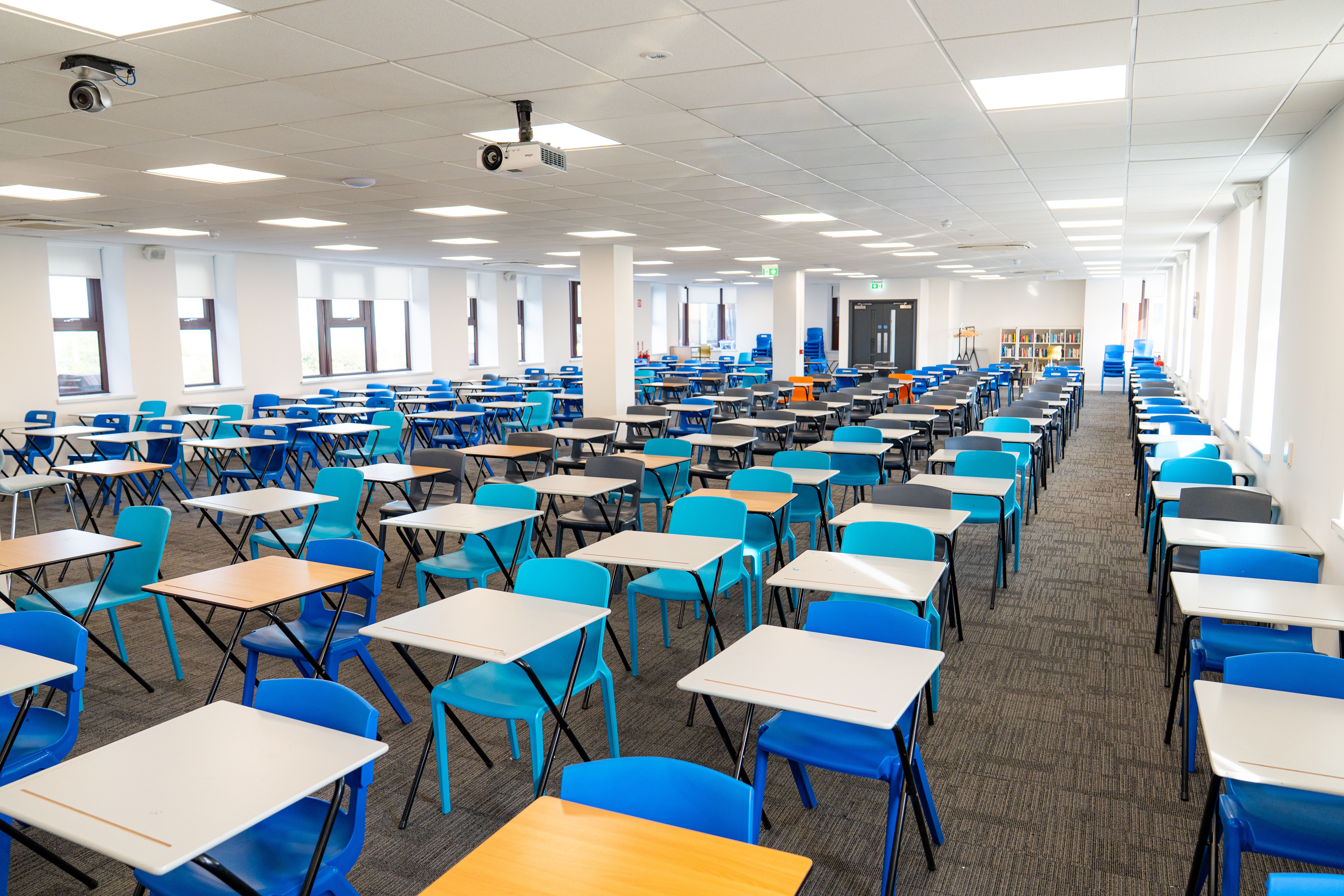 An empty classroom with rows of desks and blue chairs arranged neatly, lit by overhead lights and natural light from large windows.