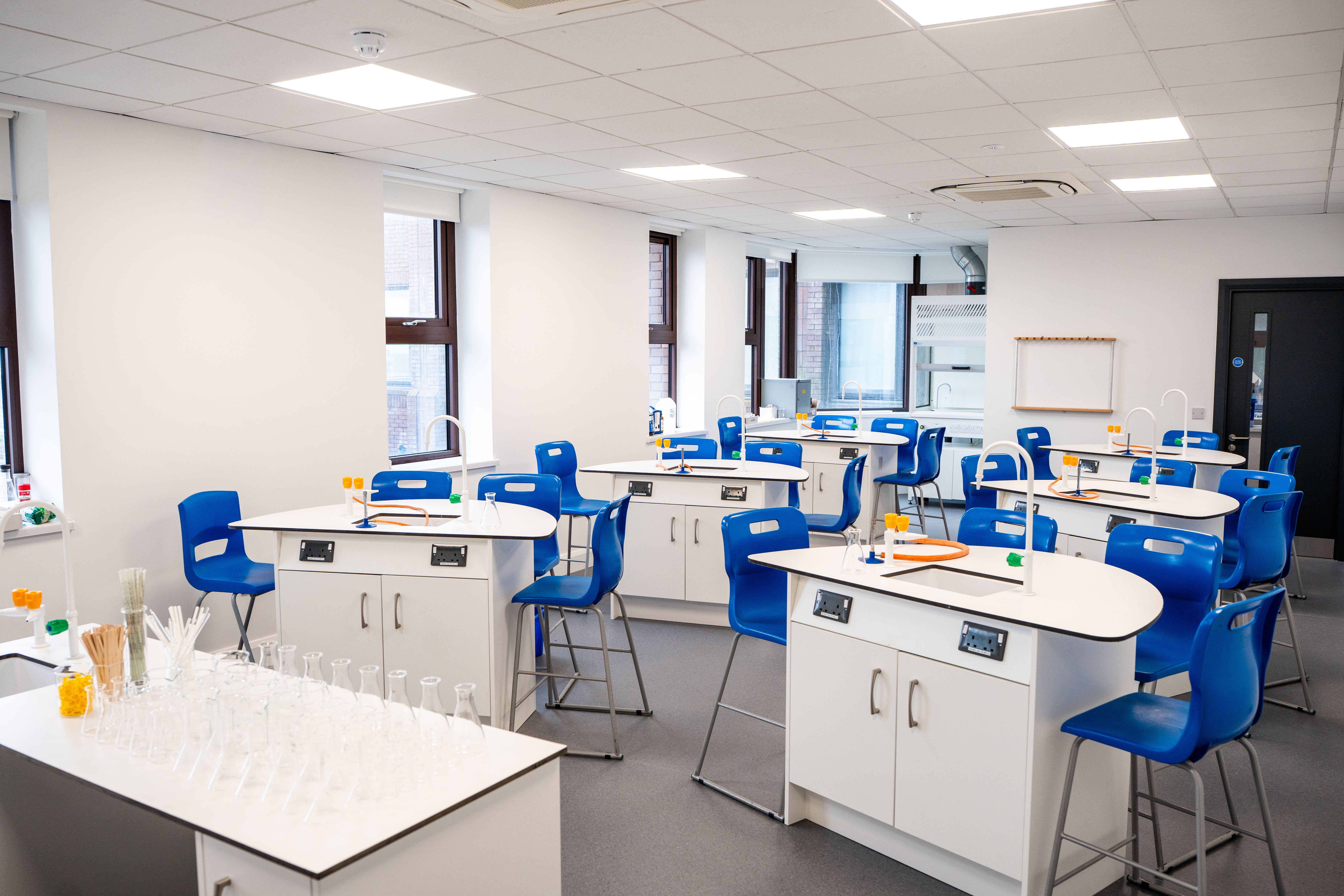 Modern science classroom with white tables, blue chairs, and lab equipment like test tubes and burners. Bright, well-lit with large windows.