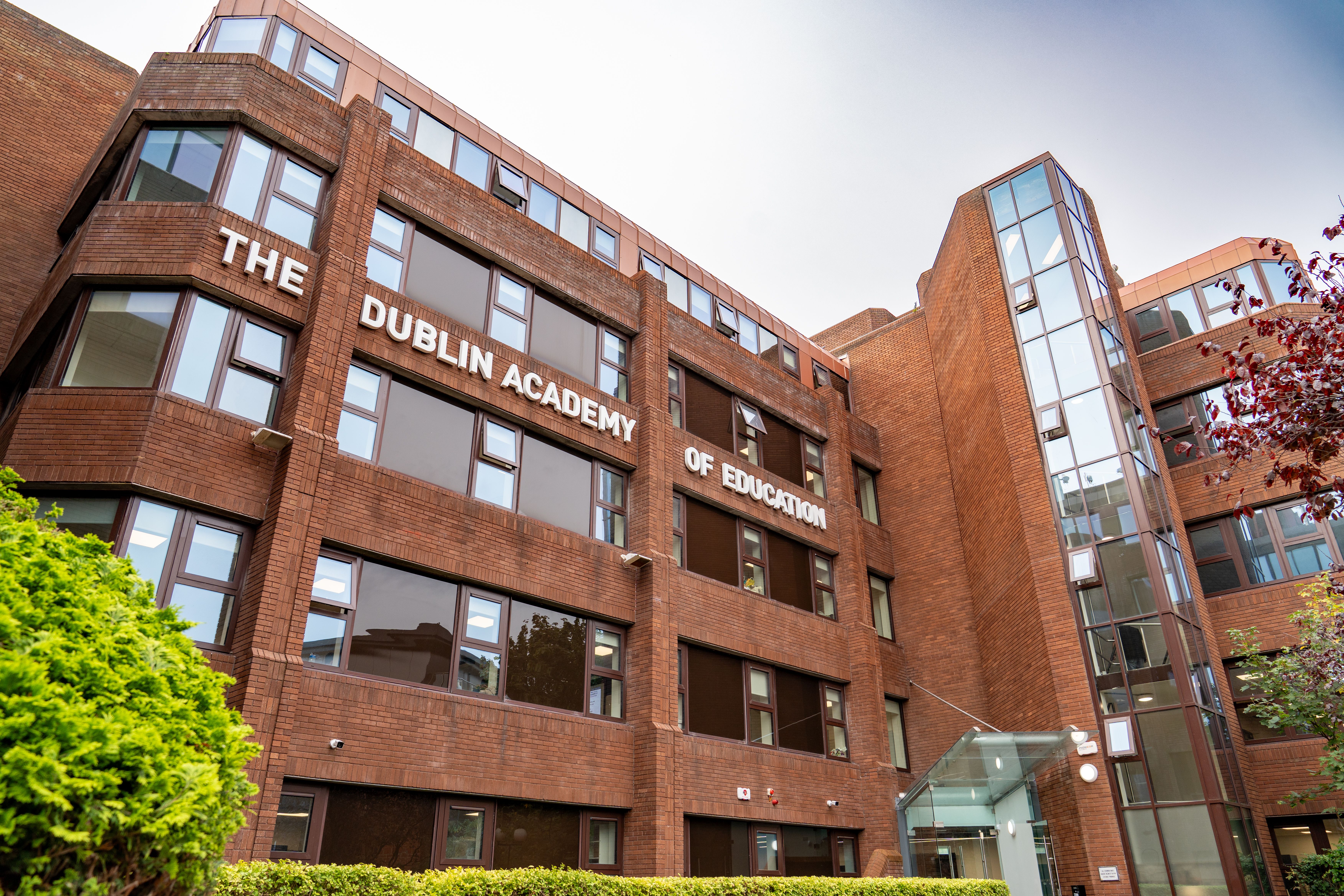 Red brick building of the Dublin Academy of Education, featuring large windows and a glass entrance, surrounded by greenery.
