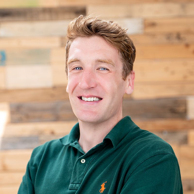 Smiling man with short hair in a green polo shirt, standing against a wooden plank background.