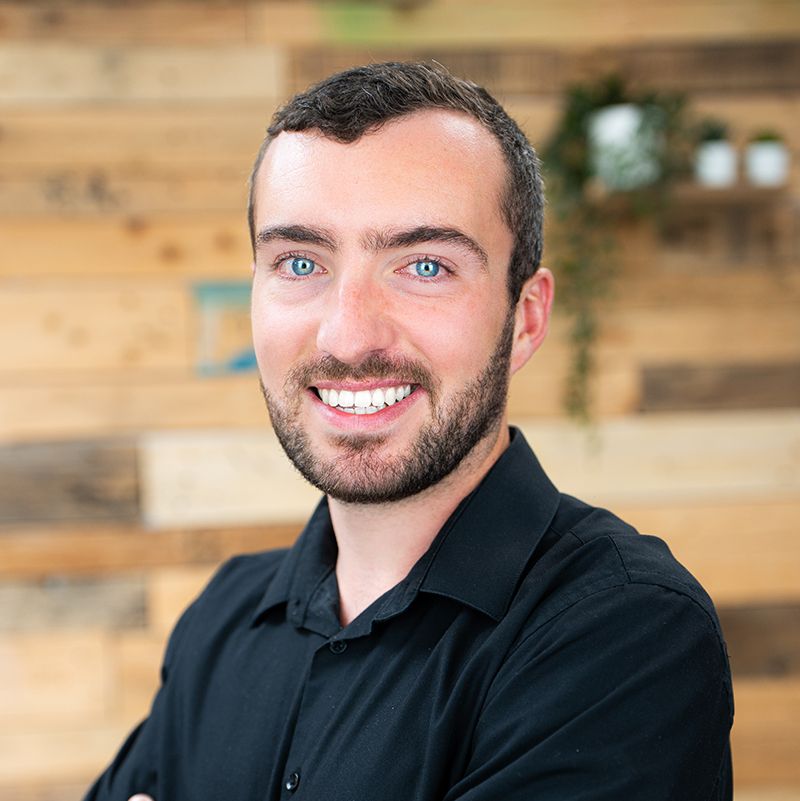 Smiling man with a beard and blue eyes, wearing a black shirt. Wooden wall with plants in the background.