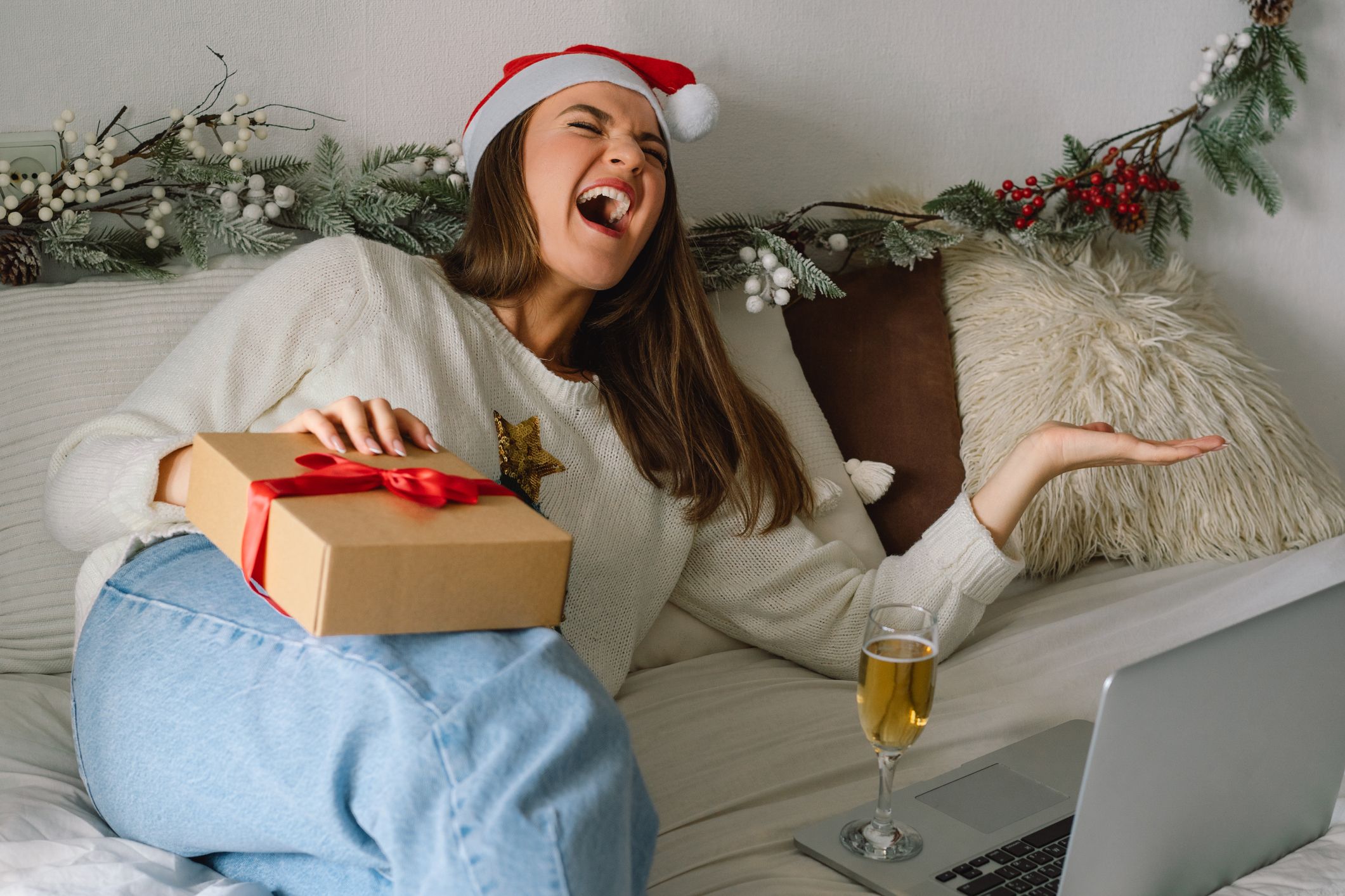woman laughing and happy at laptop with champagne, santa hat, and gift