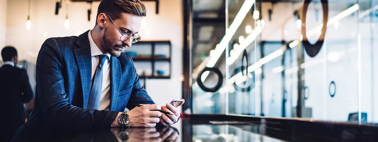 Geschäftsmann im Anzug schaut auf Smartphone an glänzendem Tisch im modernen Büro mit Glaswand und Lichtleisten.