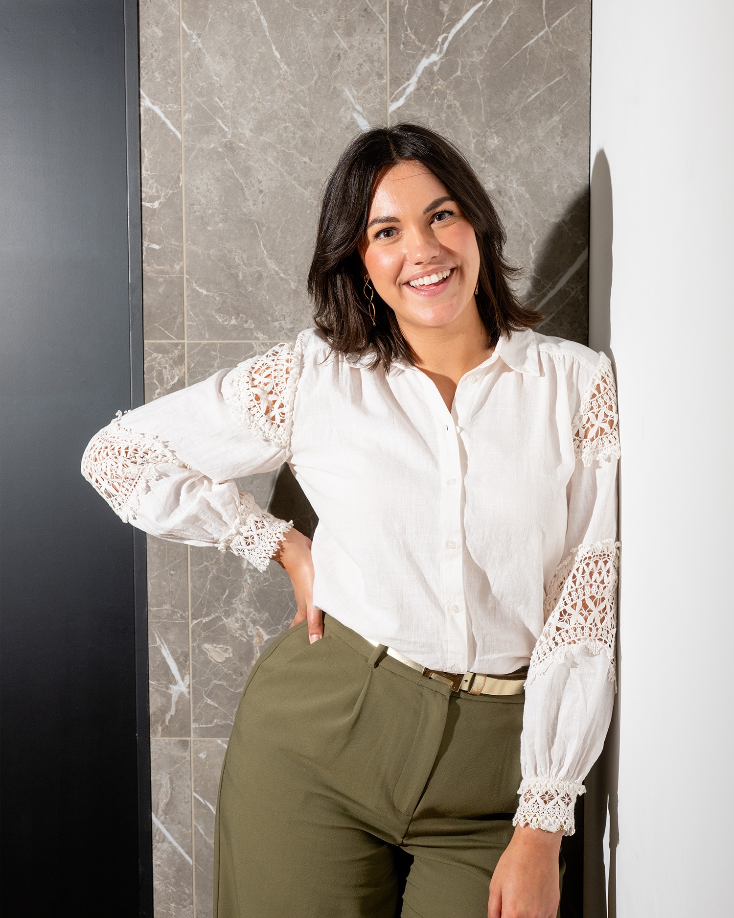 Vrouw met donker haar glimlacht in witte blouse met kant en groene broek, leunend tegen een marmeren muur.