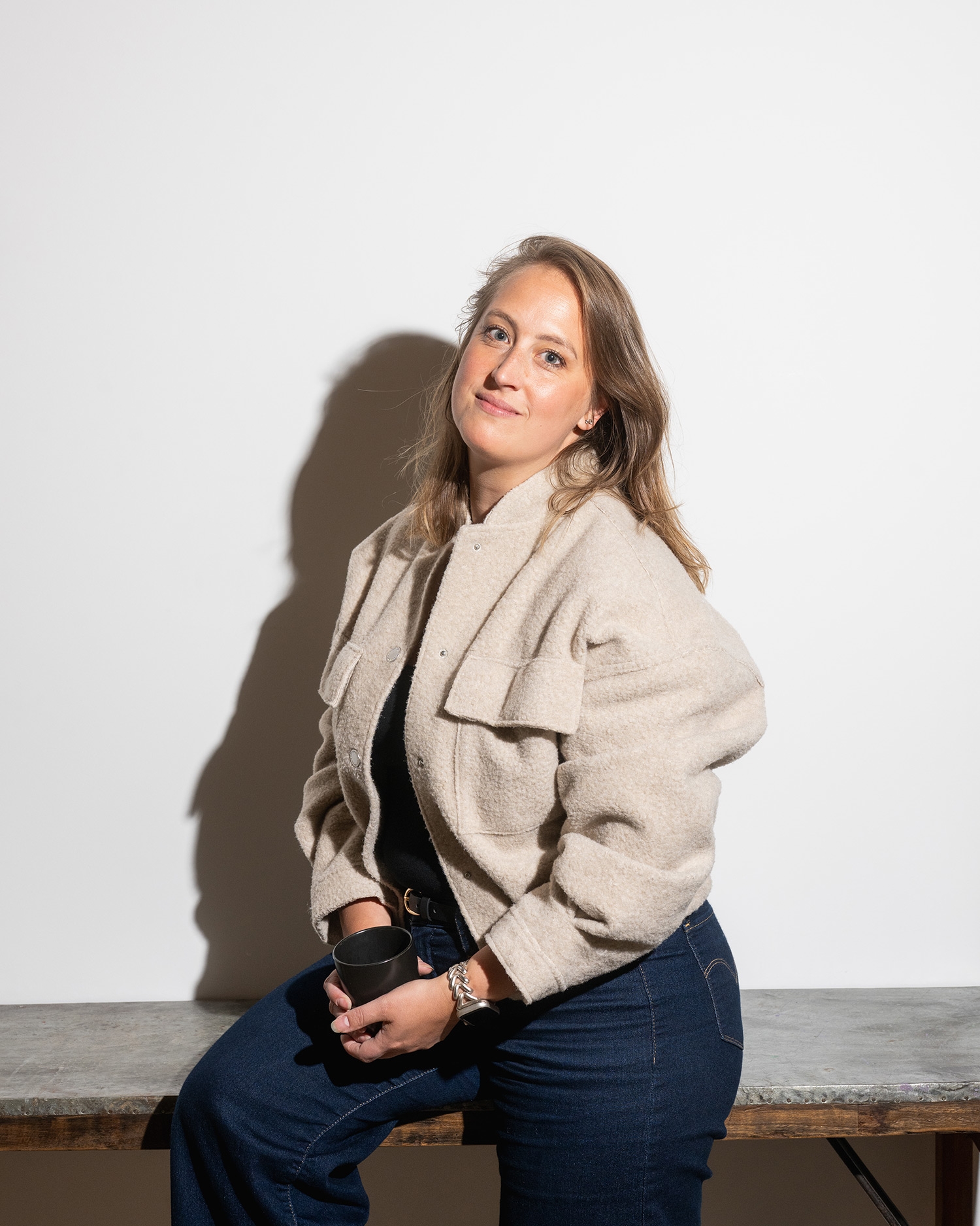 Vrouw zittend op een bank met een mok in haar hand, draagt een beige jas en blauwe broek, tegen een witte achtergrond.