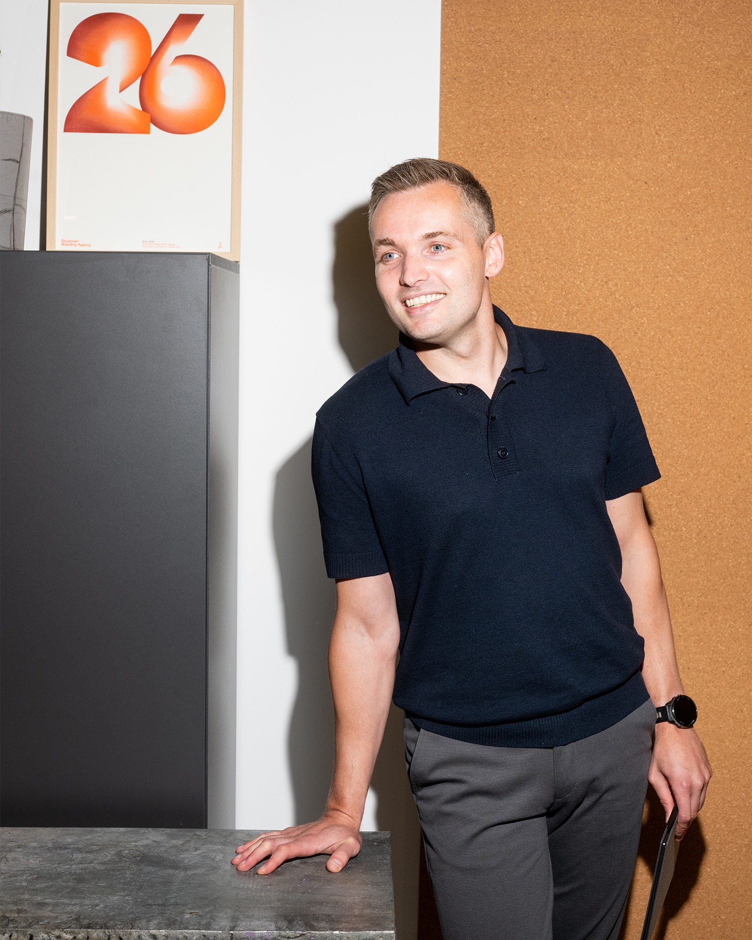 Man in donkerblauwe polo en grijze broek leunt tegen een bureau. Achtergrond met poster van het nummer 26 en kurkwand. Lachend gezicht.