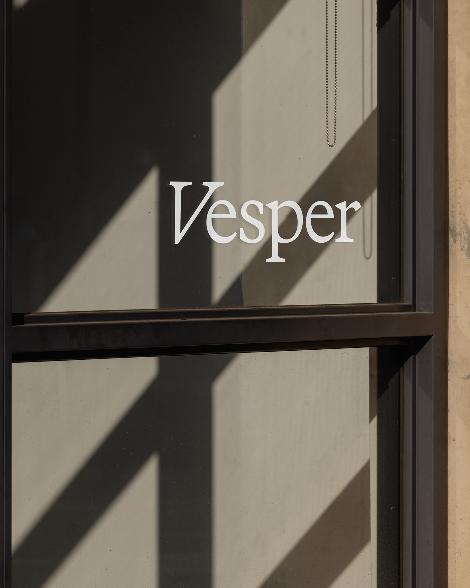 Glazen deur met het woord "Vesper" in witte letters, met schaduwen van raamkozijnen op de achtergrond.