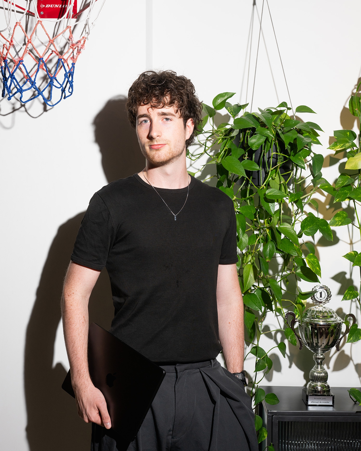 Man met krullend haar houdt een laptop vast in een kamer met planten, een bokaal en een basketbalnet op de achtergrond.