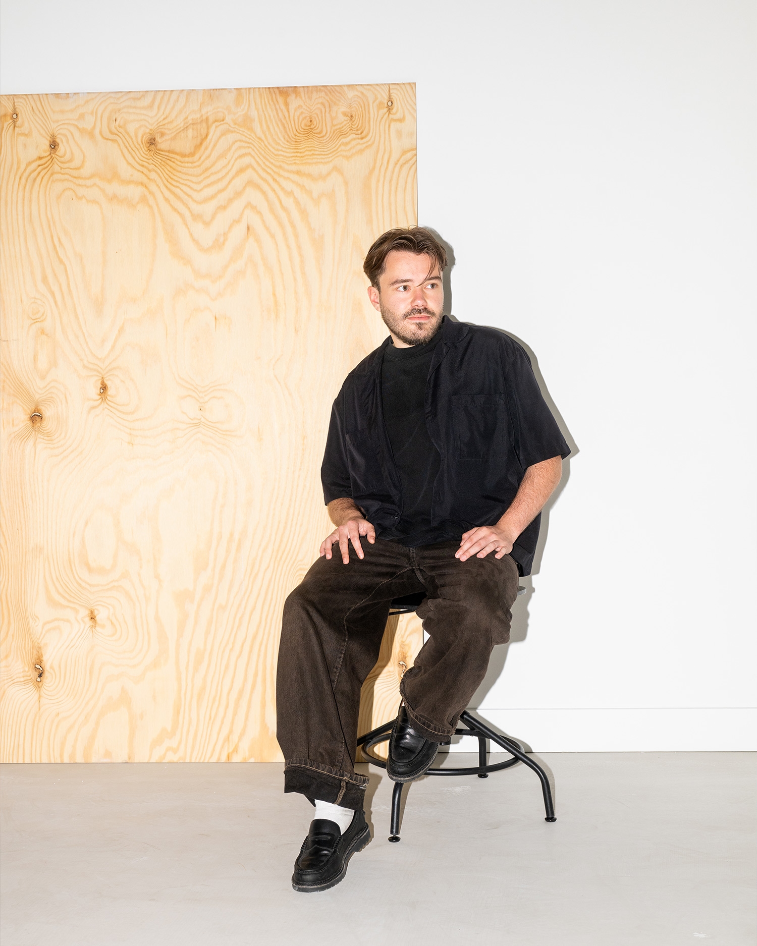 Man zit op een kruk in een minimalistisch ingerichte ruimte met een houten paneel en witte muren, gekleed in zwart shirt en bruine broek.