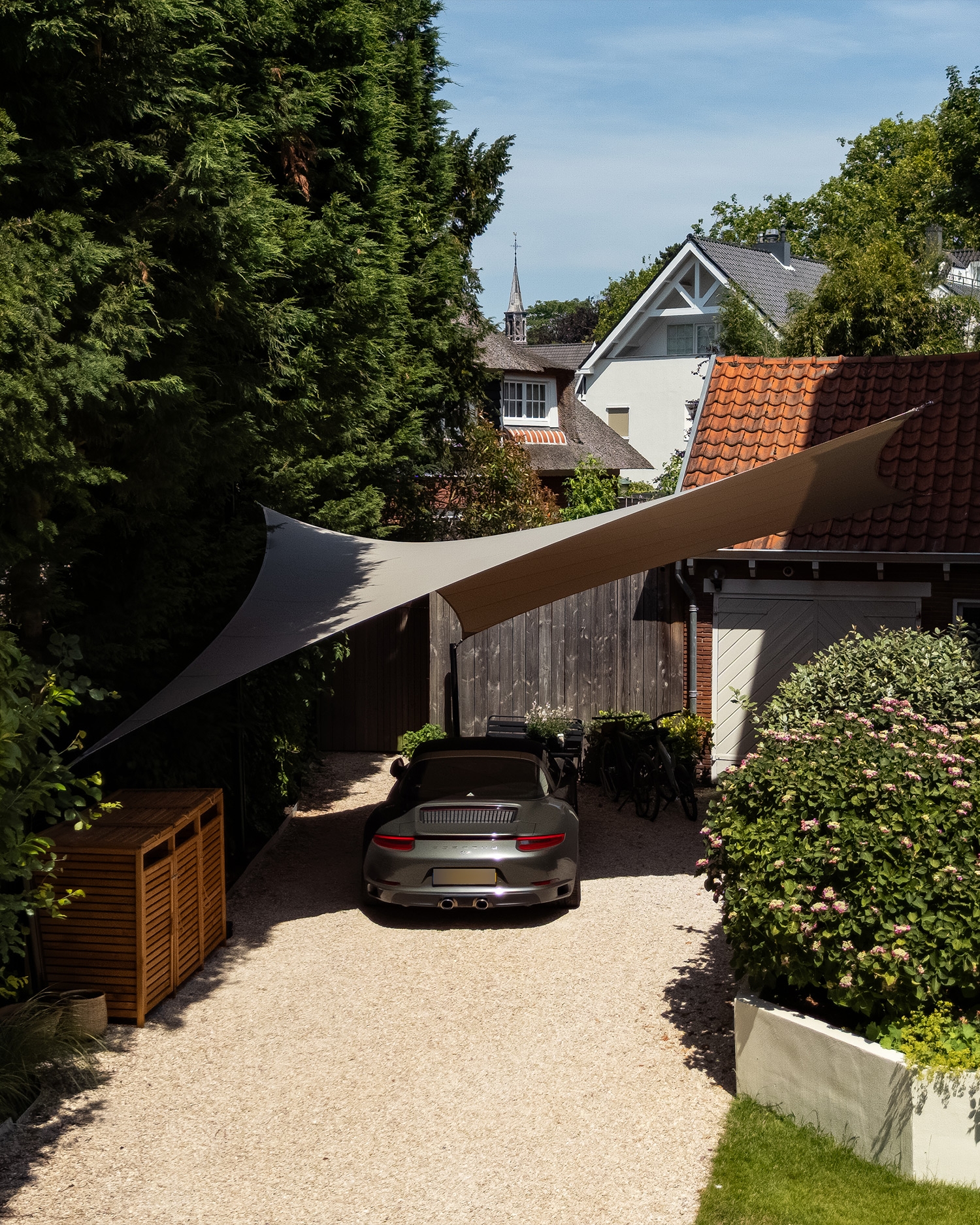 Beige Mobuline schaduwdoek boven een geparkeerde auto in een groene tuin.