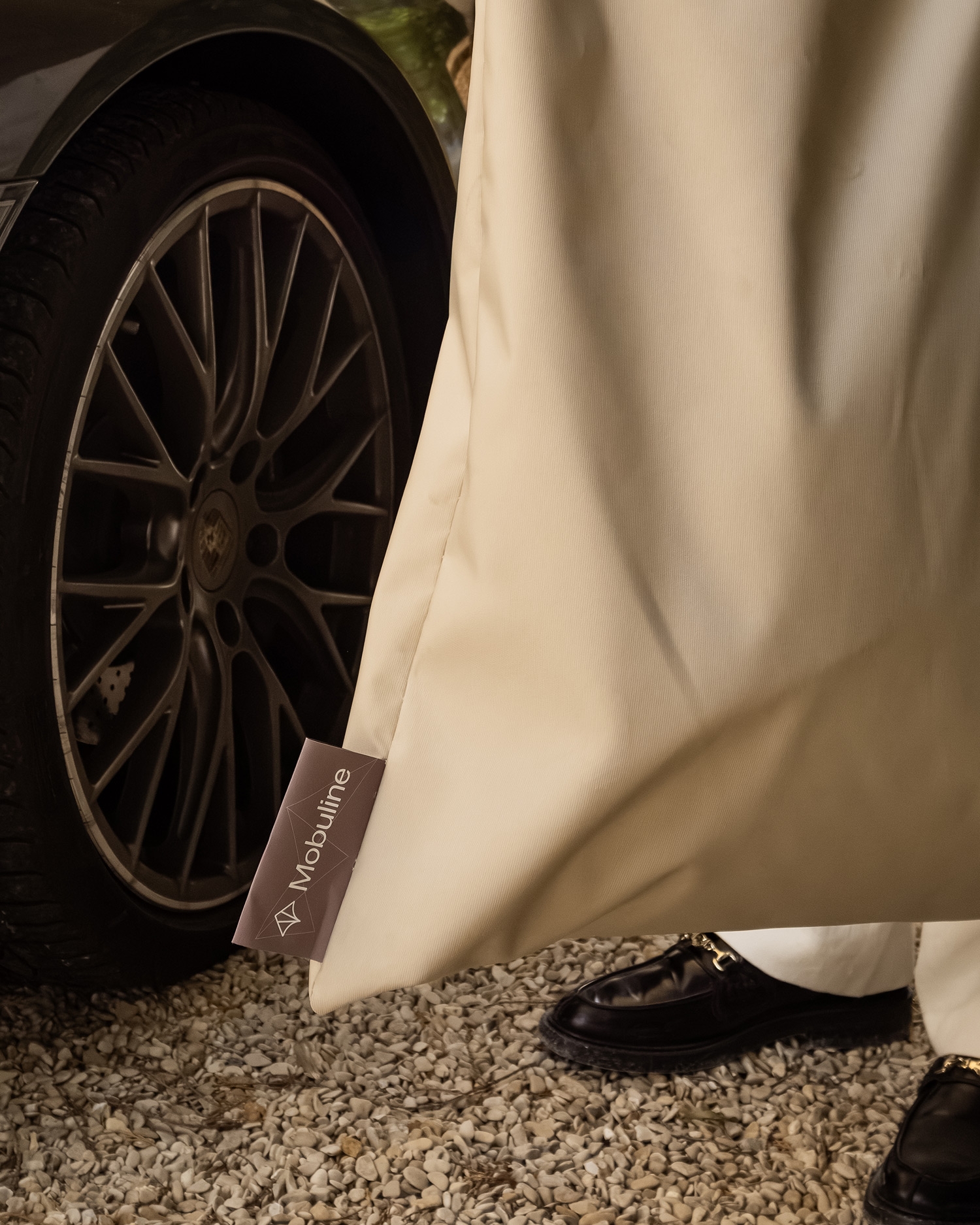 Detail van een beige Mobuline schaduwdoek naast het wiel van een donkere auto.