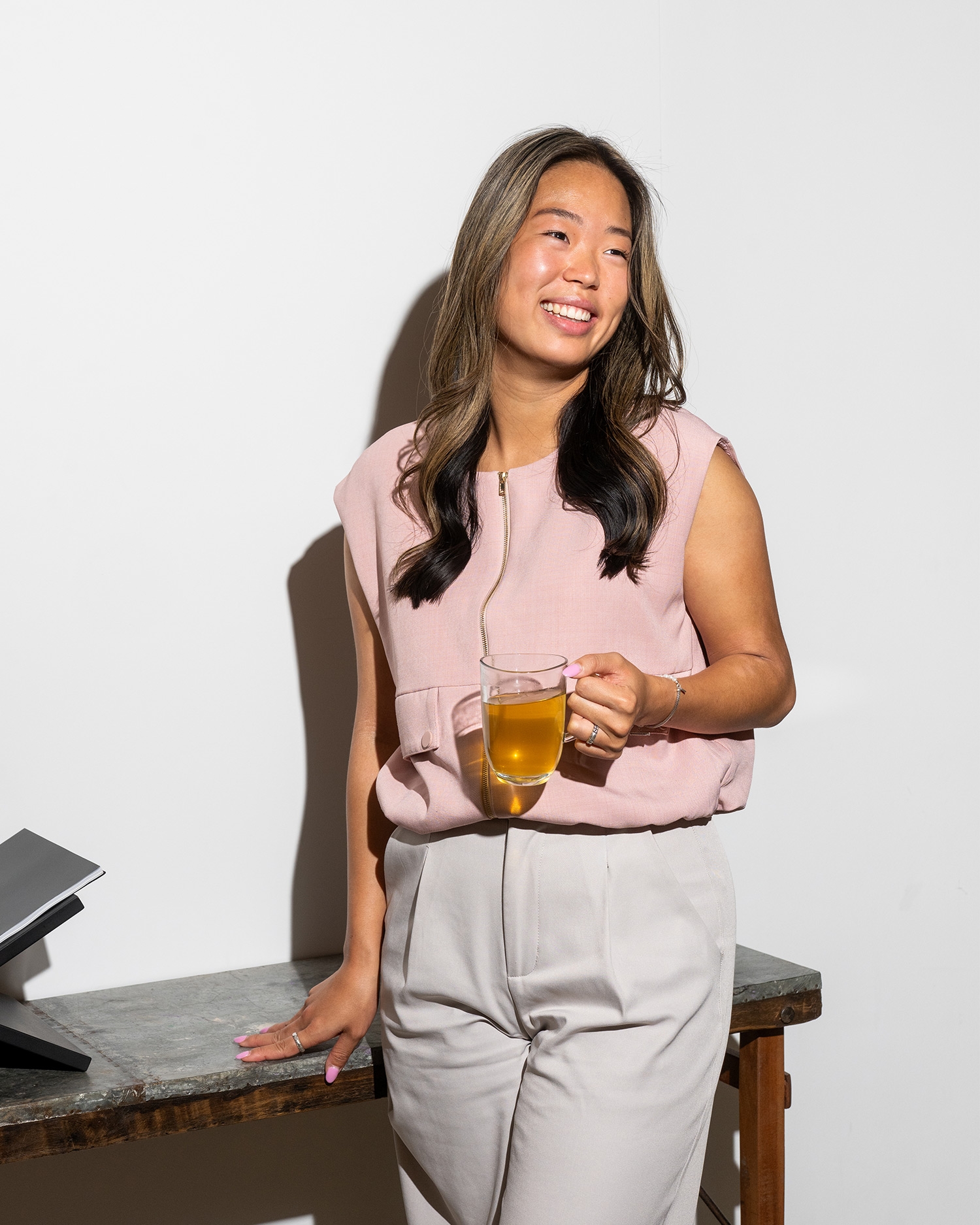 Maria in een roze blouse en grijze broek leunt lachend tegen een tafel, met een glas thee in haar hand.