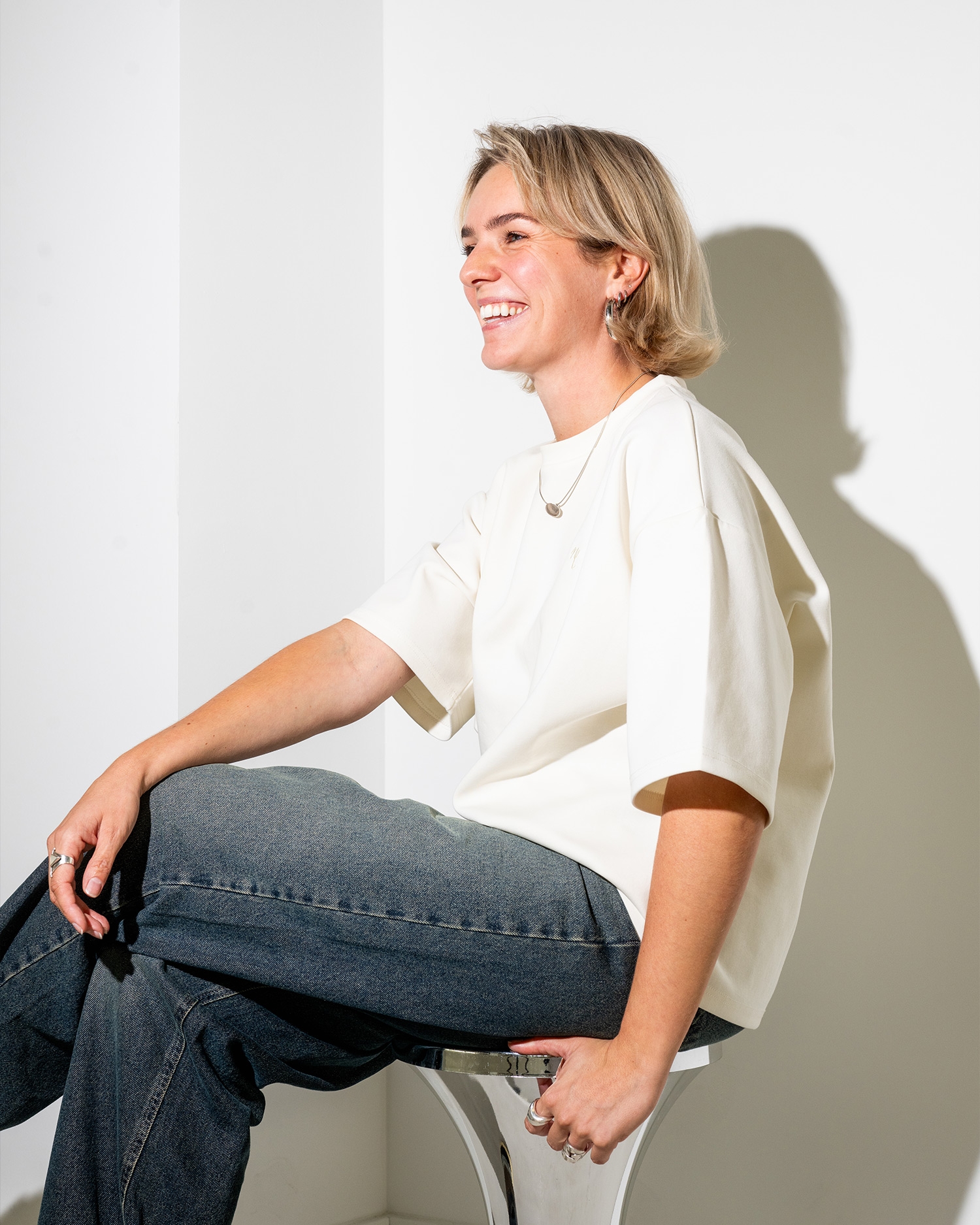 Persoon met kort blond haar lacht, zittend op een stoel. Ze draagt een wit T-shirt en een spijkerbroek. Schaduw op witte muur.