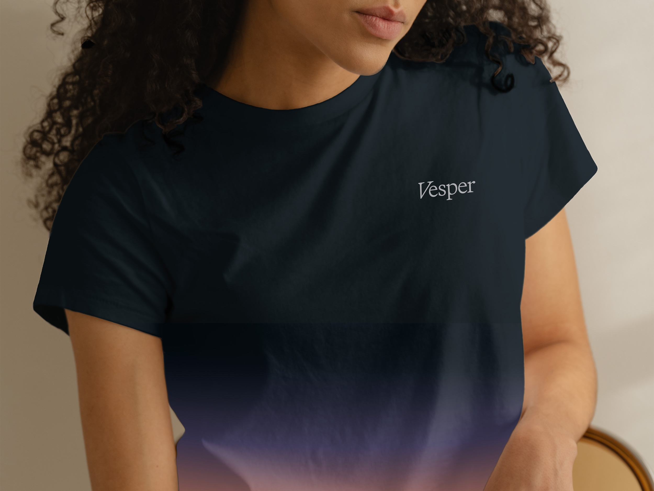 Vrouw met donkere krullen draagt een donker shirt met de tekst "Vesper" aan de voorkant, gedeeltelijk in beeld, tegen een neutrale achtergrond.