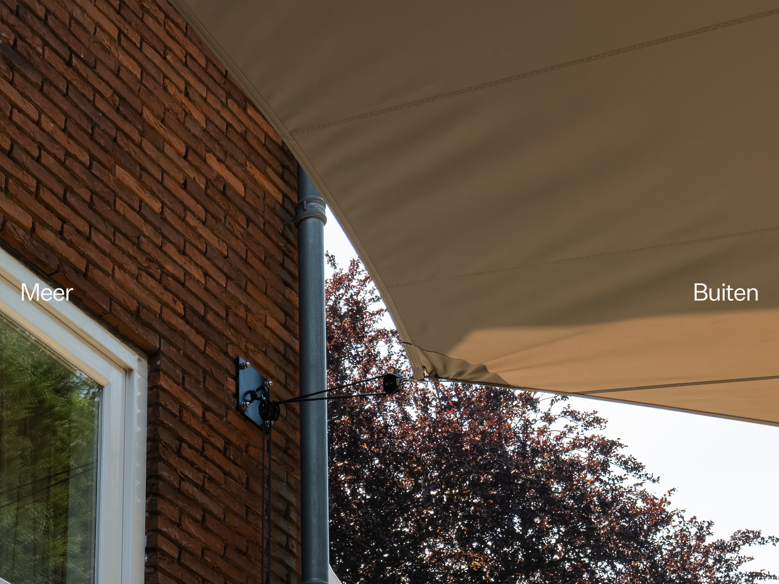 Detail van een beige schaduwdoek aan een bakstenen woning met de woorden Meer en Buiten.