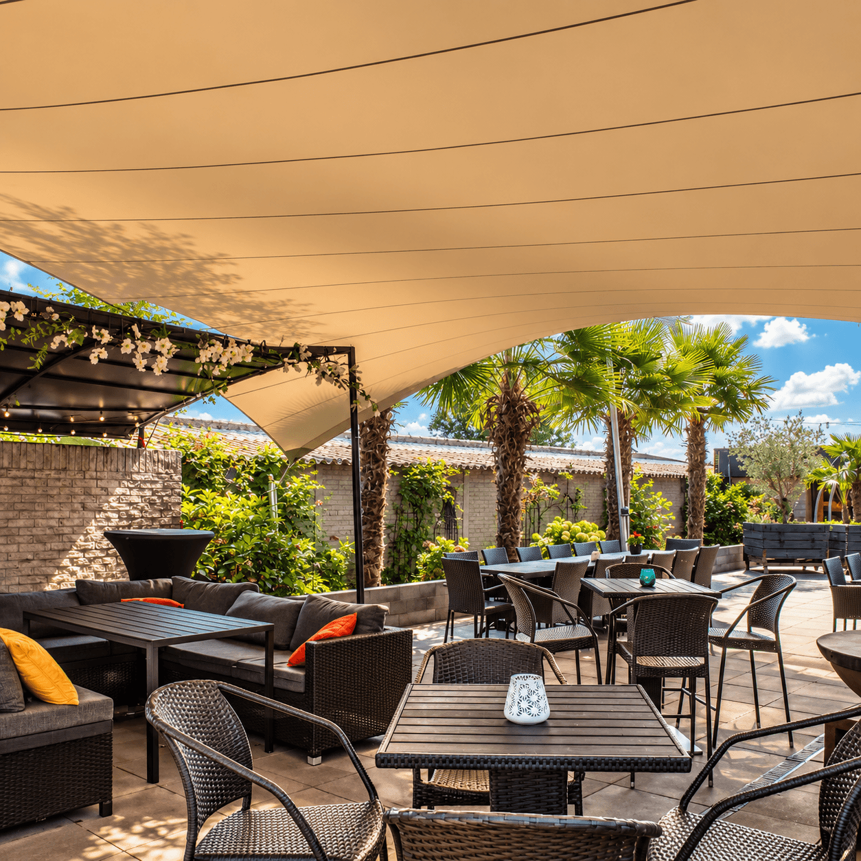 Beige maatwerk zonnedoek boven een terras aan een karakteristiek stenen gebouw, gespannen tussen gevel en masten.
