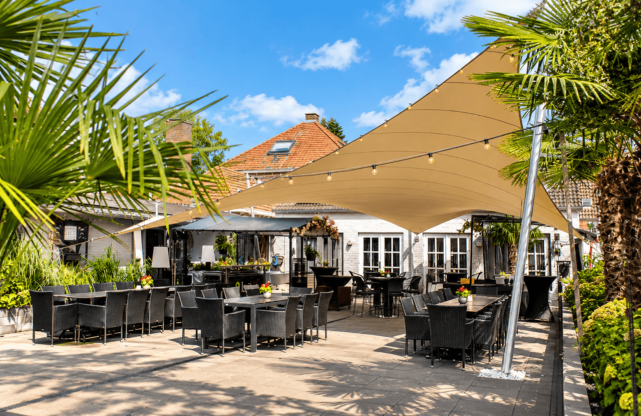 Ruime maatwerk membraanoverkapping boven een ingericht horecaterras, omringd door palmbomen en beplanting.