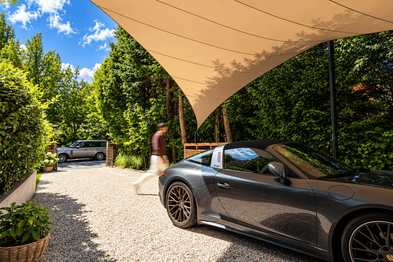 Beigefarbenes Architektur-Segeldach von Mobuline als maßgefertigter Design-Carport über einem grauen Sportwagen auf einer von grünen Hecken umgebenen Einfahrt.