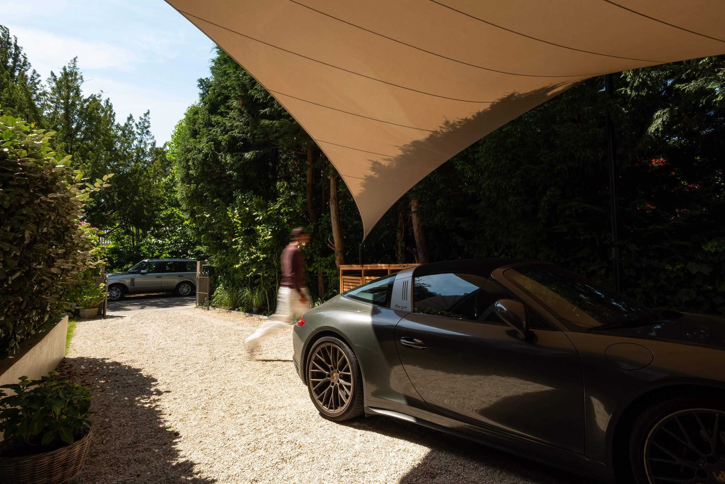 A sleek black convertible is parked under a large canopy, with a blurred figure walking past and a car in the background surrounded by lush trees.