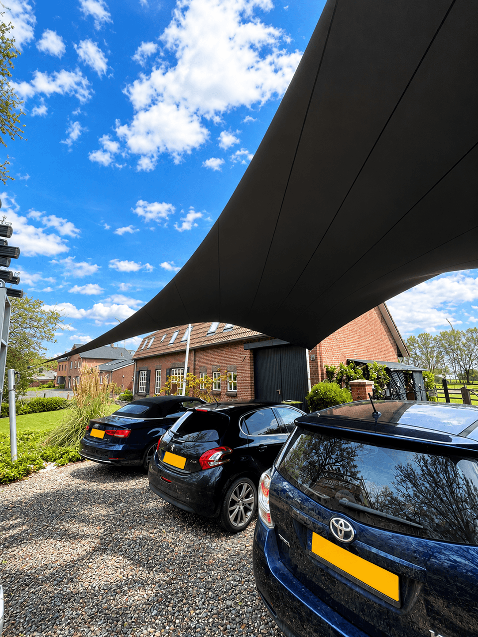 Zwarte design carport van gespannen textiel boven drie geparkeerde auto’s bij een landelijke woning.