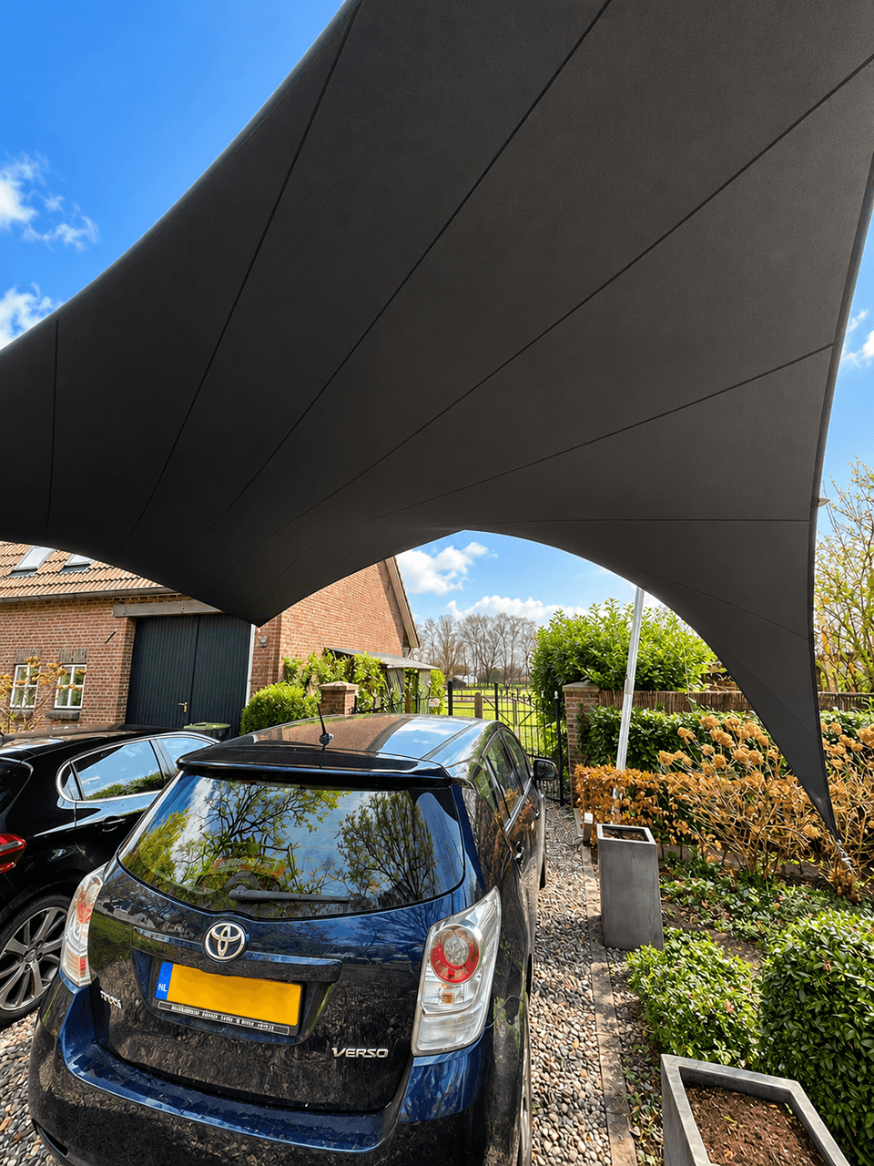 Zwart maatwerk schaduwdoek als carport boven geparkeerde auto’s bij een woning.