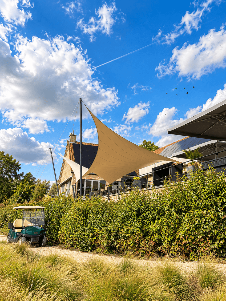 Groot bruin maatwerk zonnedoek boven het terras van een golfclub, gespannen tussen hoge masten.