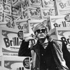 black and white photograph of Andy Warhol wearing sunglasses in front of a stack of Brillo boxes