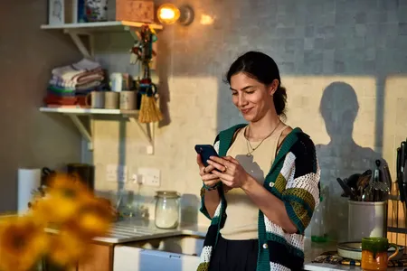 woman smiling at phone standing in sunlit kitchen