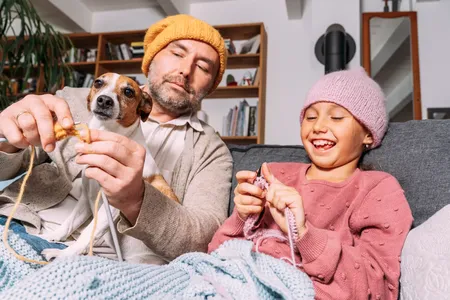 father and daughter happily knitting hats on the sofa with their dog